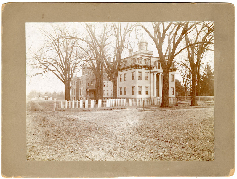 Photograph of white square building with white fence and trees in foreground