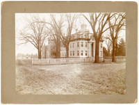 Photograph of white square building with white fence and trees in foreground
