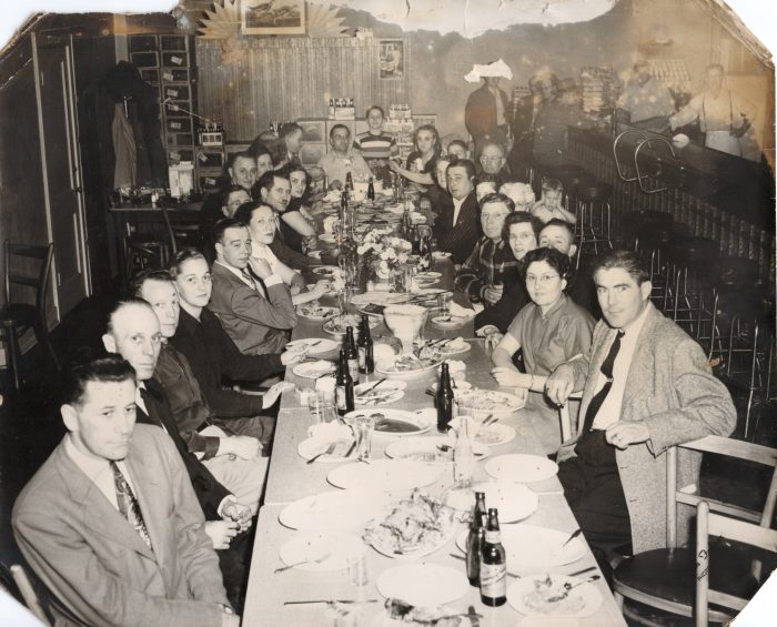 A black-and-white photograph of thirty people seated around a long table. There are plates and bottles of beer on the table.